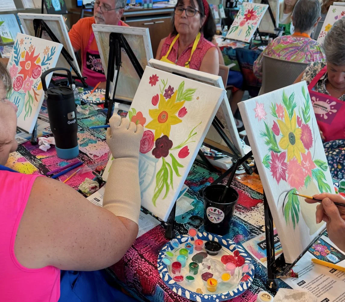 People participating in a group painting class, creating floral artwork on canvases set on small easels.