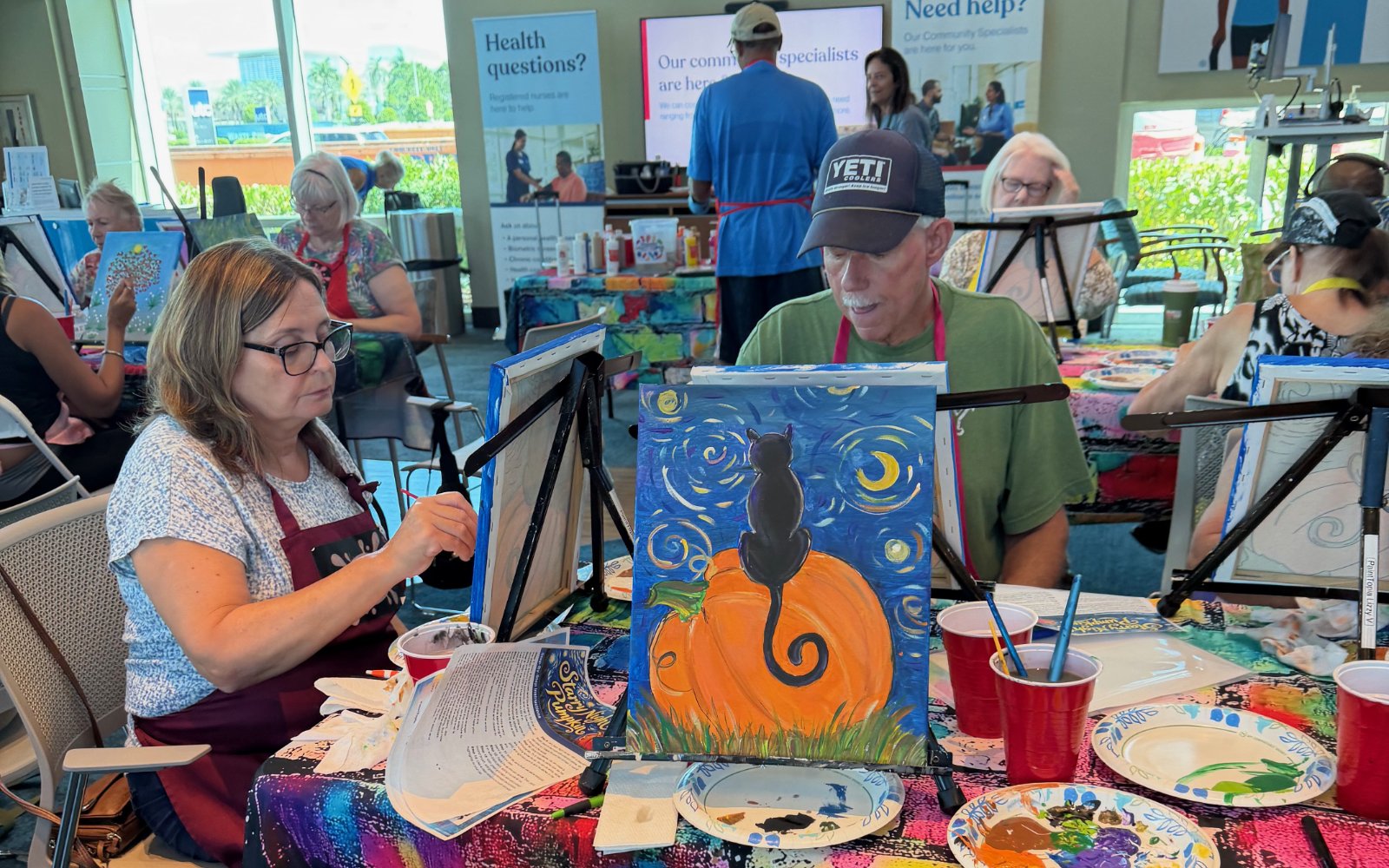 People participating in a group painting activity at a community event, with a canvas of a black cat sitting on a pumpkin in the foreground, and a colorful table with art supplies.
