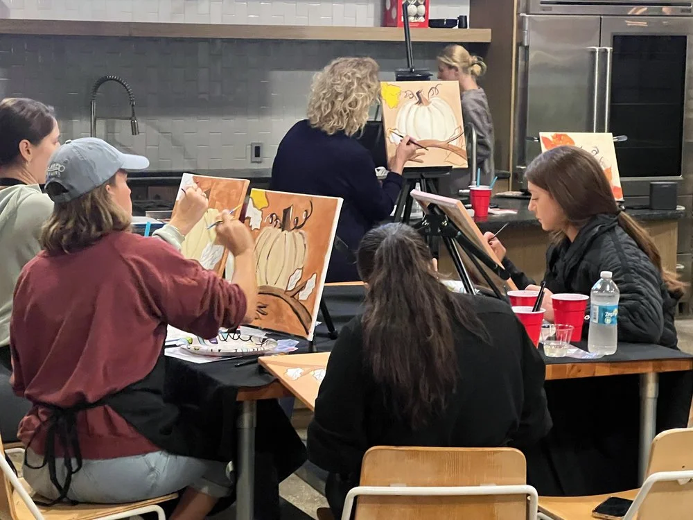 People painting fall-themed pumpkins on canvases at a group art class in a kitchen setting.