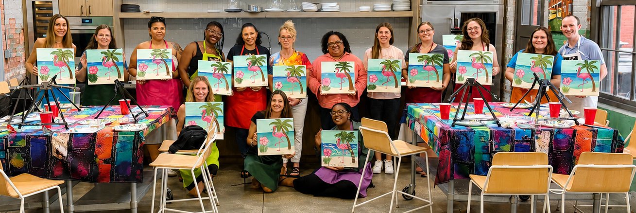 A group of diverse women in an art class holding up paintings of pink flamingos on a tropical background, in a colorful classroom.