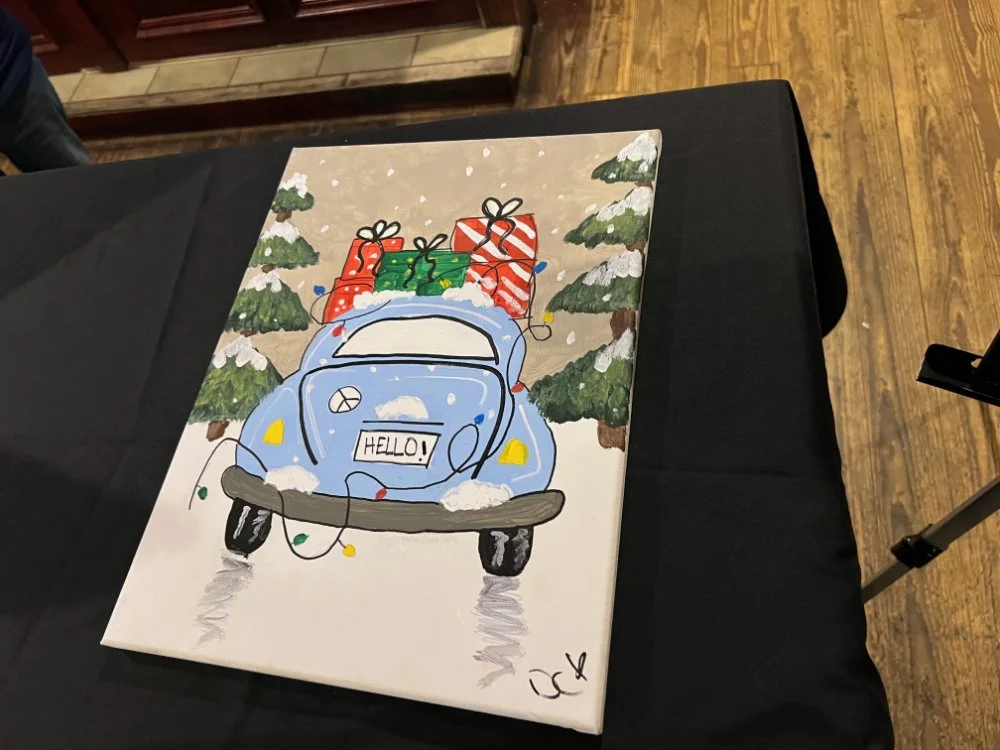 Painting of a blue vintage car with snow, topped with Christmas gifts, flanked by snow-covered Christmas trees, on a black table.