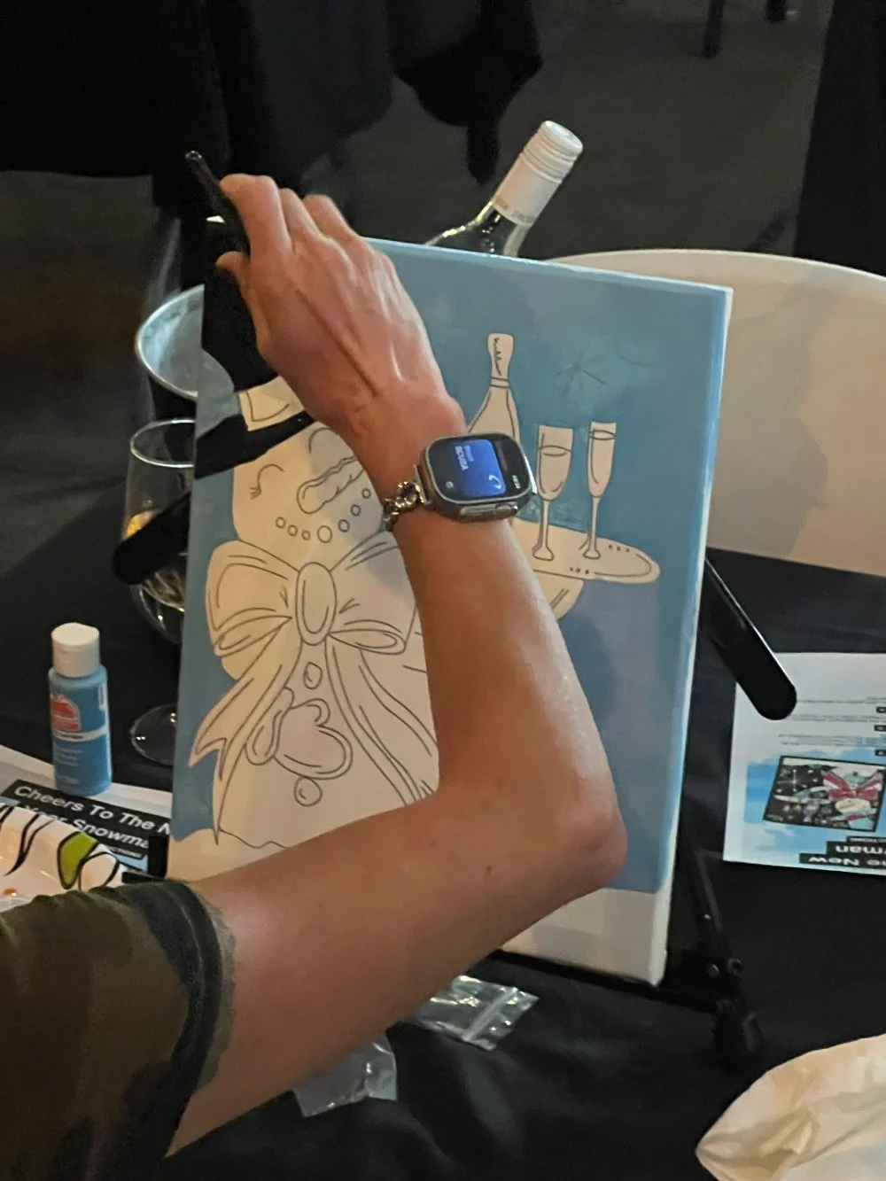A person coloring a festive holiday-themed drawing of a bow and holly leaves on a blue background, with several glasses and a bottle of white wine visible on the table.