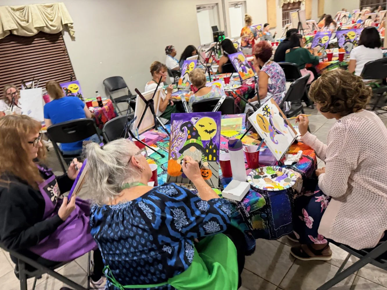 Group of women participating in a painting class at a Halloween-themed event, with spooky decorations and colorful paintings on easels.