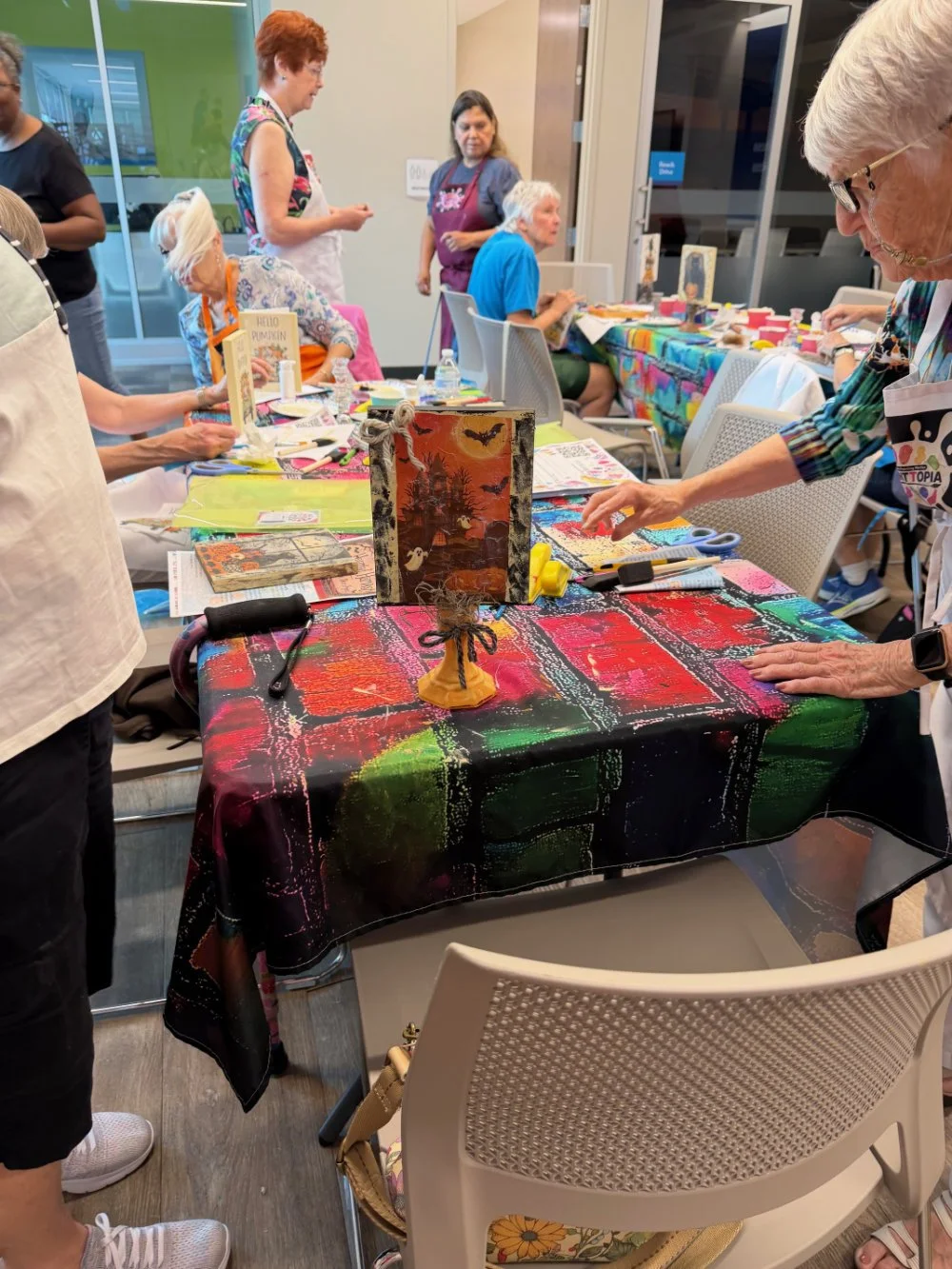 People participating in a craft workshop, working on decorated tables with art supplies and a Halloween-themed centerpiece.