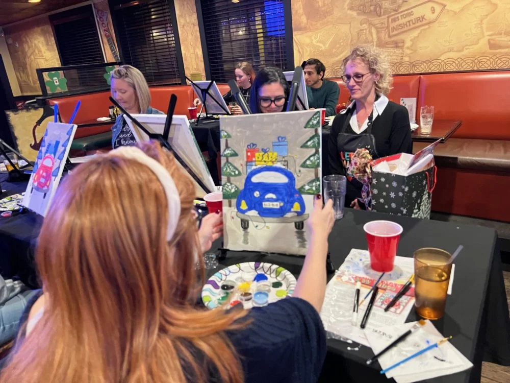 A group of people participating in a painting on canvas activity at a restaurant or bar, with festive decorations and gift bags visible.