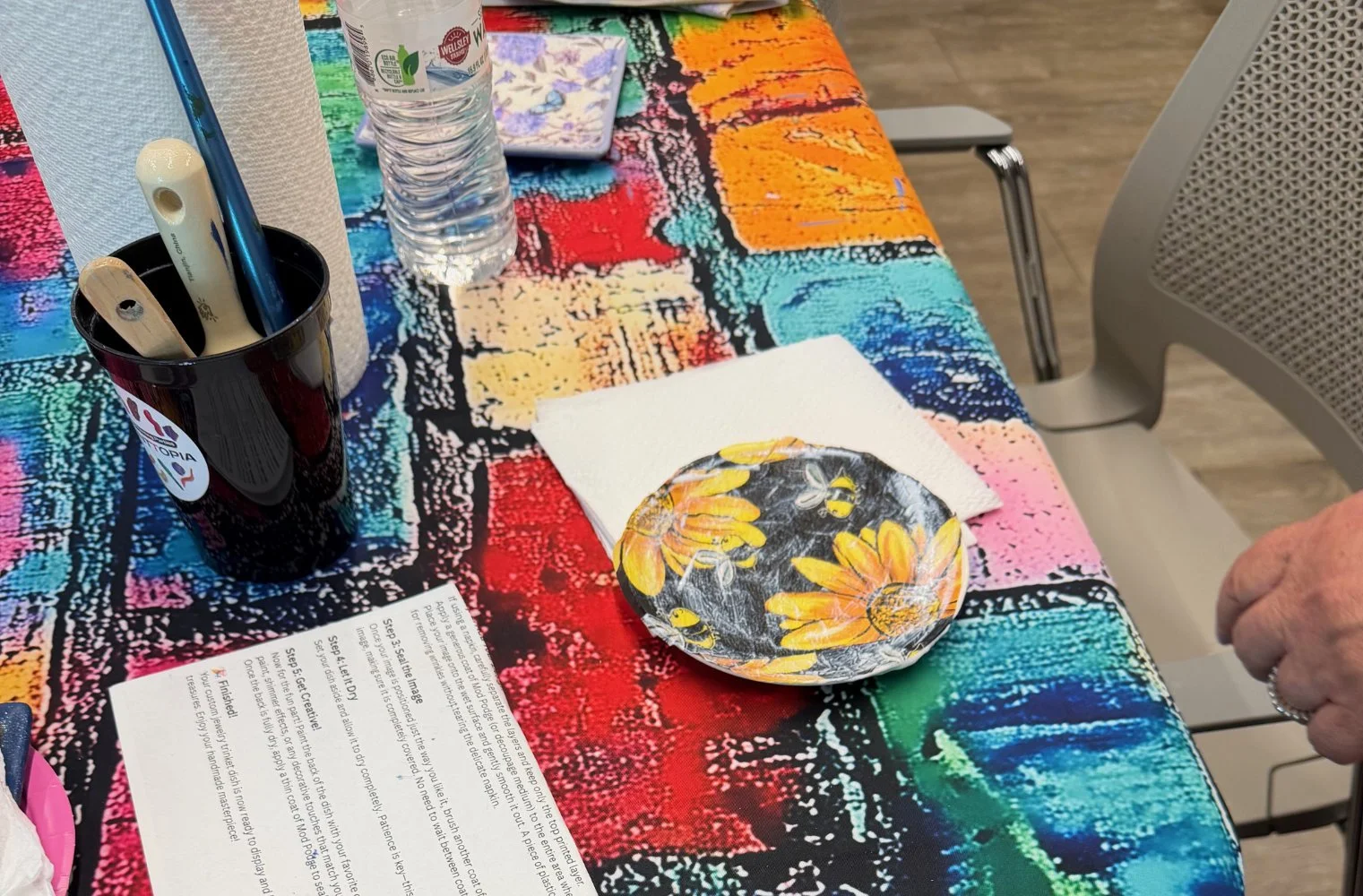 A colorful tablecloth with a floral plate, a paper napkin, a bottle of water, a black cup holding pens and a paintbrush, and a piece of paper.