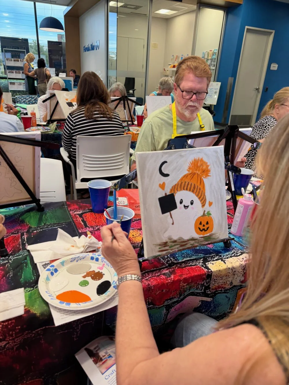 People participating in a paint and sip art class, creating Halloween-themed artwork featuring a cute ghost with a pumpkin, surrounded by hearts, on a colorful table.