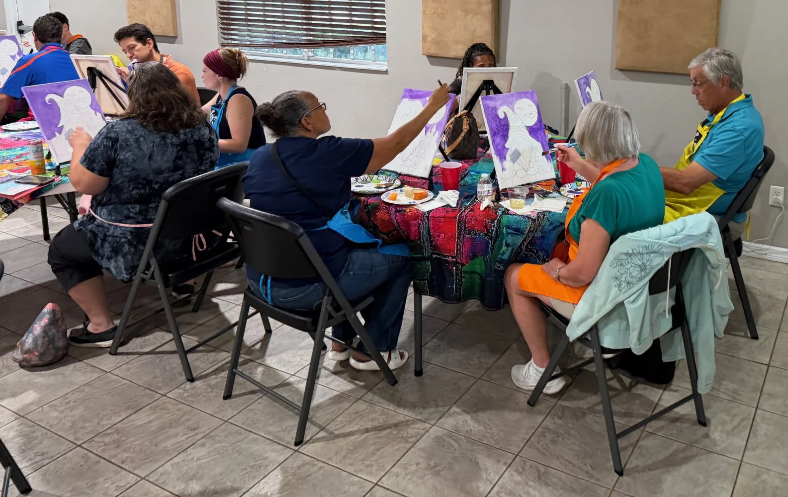 Group of people painting during an art class in a room with colorful decor, sitting around a table with painting supplies.