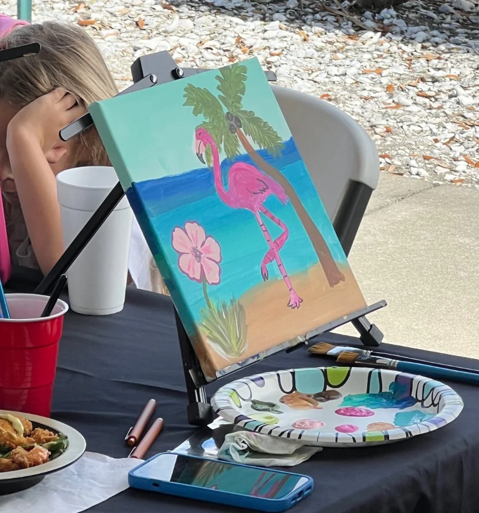 A young girl resting her head on her hand at an outdoor painting station with a canvas of a pink flamingo, palm trees, and a flower, along with painting supplies and a plate of food on the table.