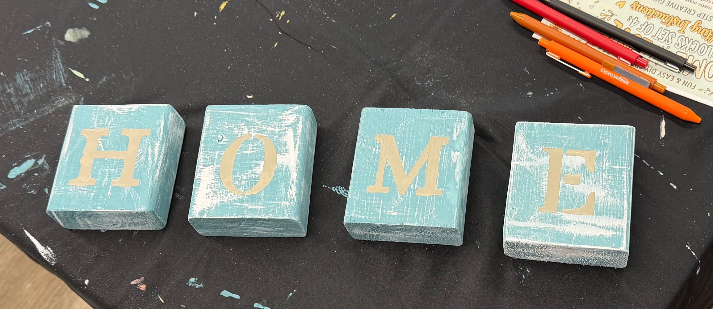 Four painted wooden blocks spelling out the word "HOME" on a black surface with art supplies nearby.