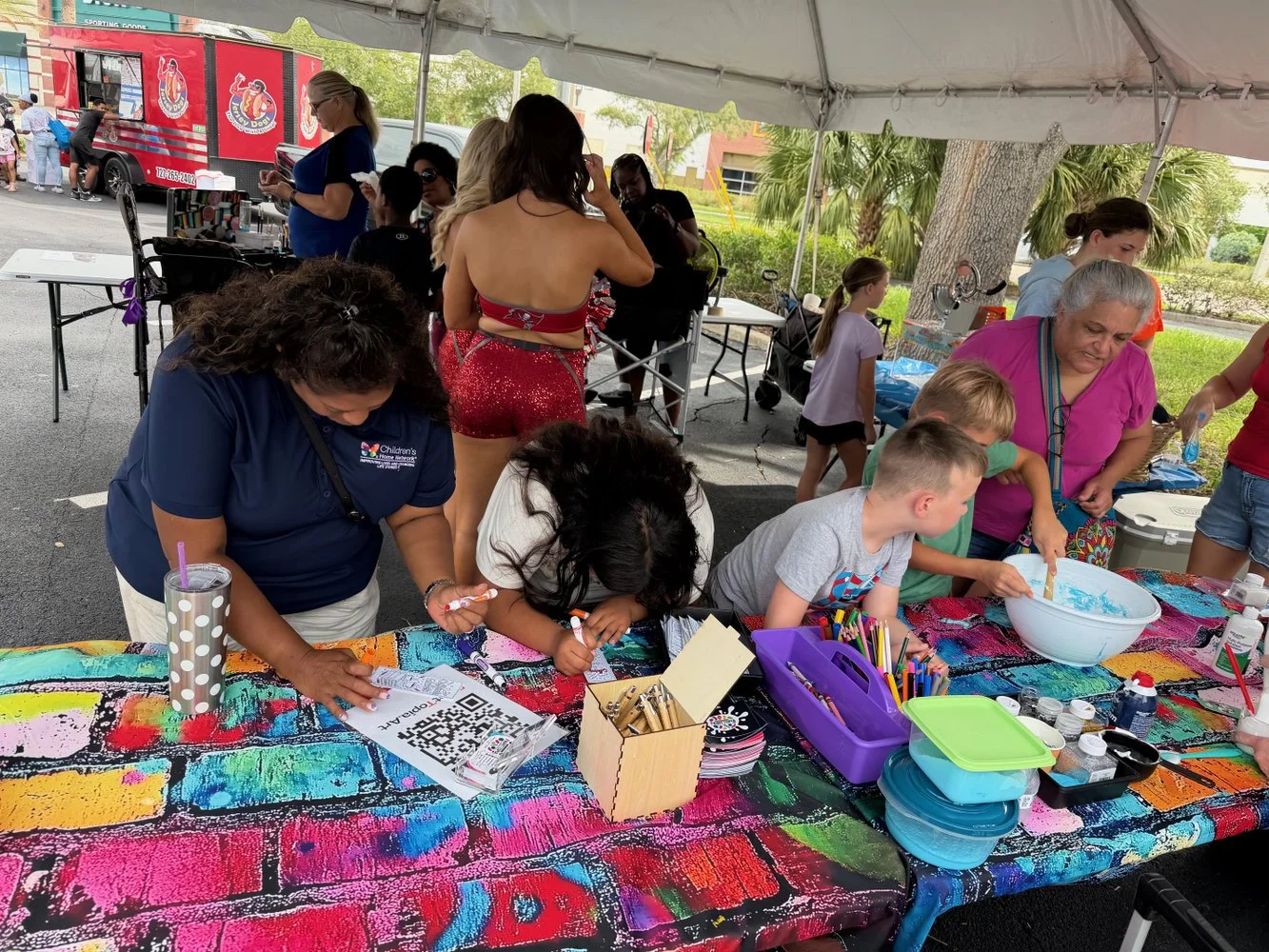 Children and adults participating in a craft activity under a tent at an outdoor event, with tables covered in colorful graffiti-style art, art supplies, and a food truck in the background.