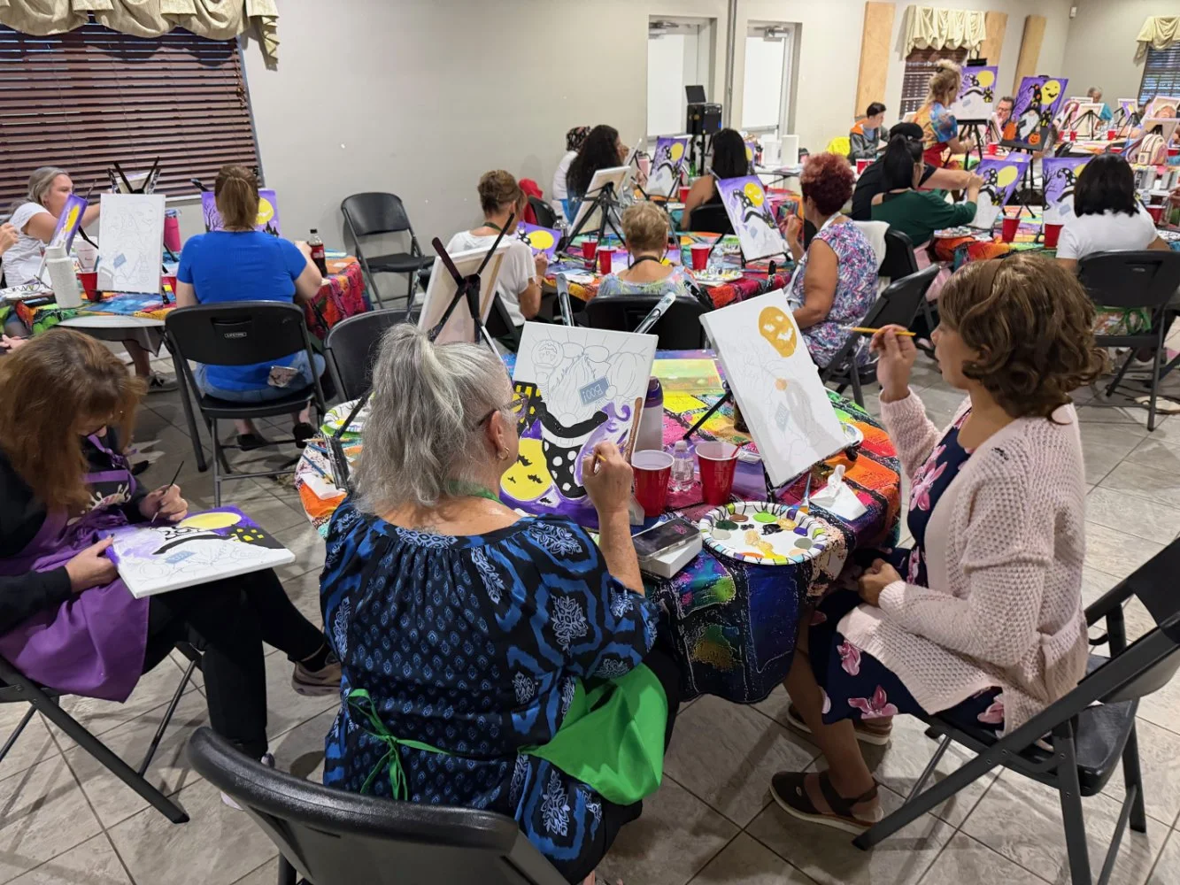 People participating in a group painting class, seated at tables with art supplies, working on Halloween-themed artwork with pumpkins and ghosts.