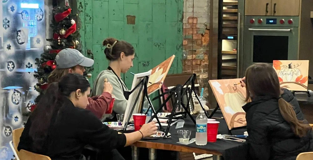 Five women painting on canvases sit around a table in a festive, decorated room with a Christmas tree and holiday decorations in the background.