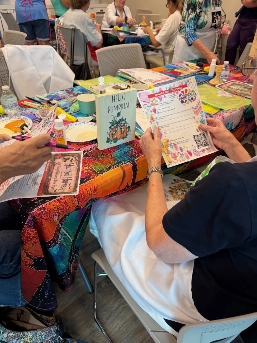Group of people at a craft workshop, with colorful supplies on the table, including glue, paper, and paint, participating in a pumpkin-themed craft activity.