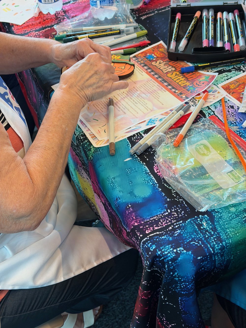 Close-up of an elderly person creating Halloween decorations on a colorful, tie-dye tablecloth with markers, stickers, and printed paper decorations around.