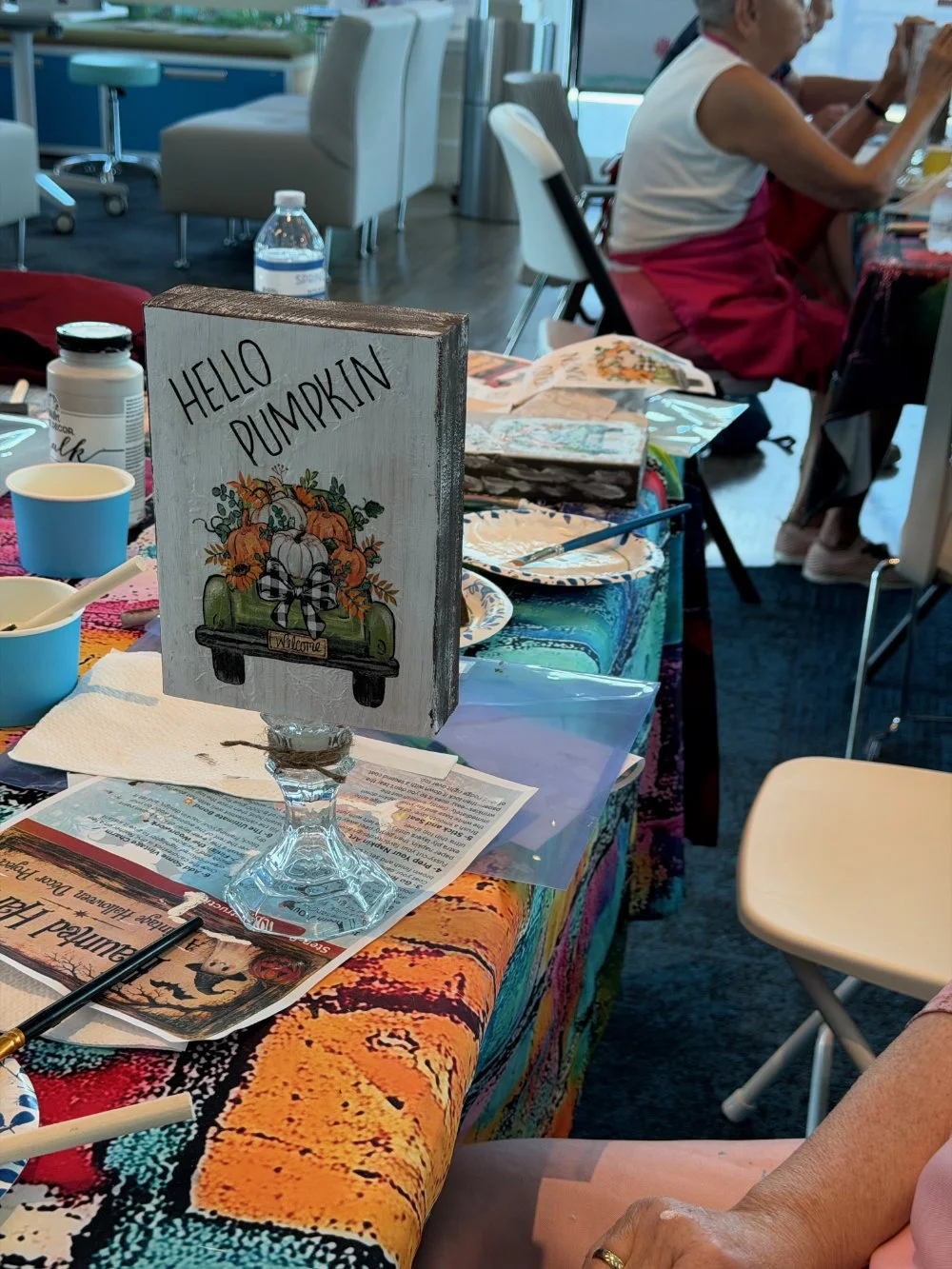A table decorated with a colorful tablecloth holds a sign that says 'HELLO PUMPKIN' with an illustration of a truck carrying pumpkins. The table has paper plates, bowls, a bottle of water, and various papers. In the background, an older woman sitting
