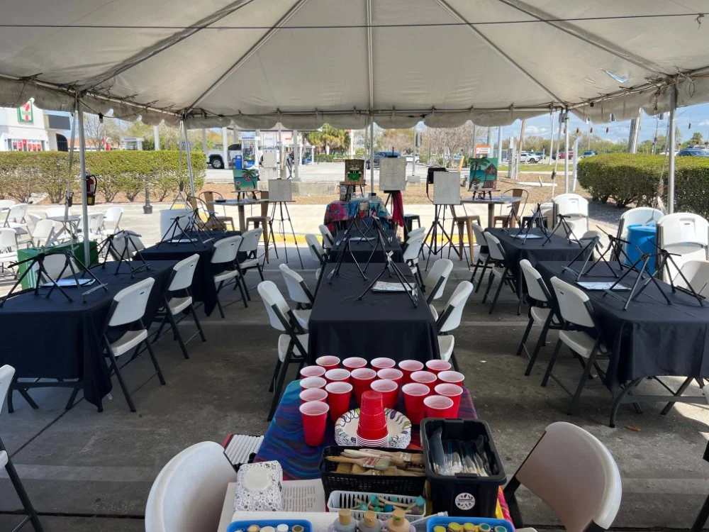 Empty outdoor event setup under a large tent with black tables, white chairs, red cups on a table in the foreground, and painting canvases on easels in the background.