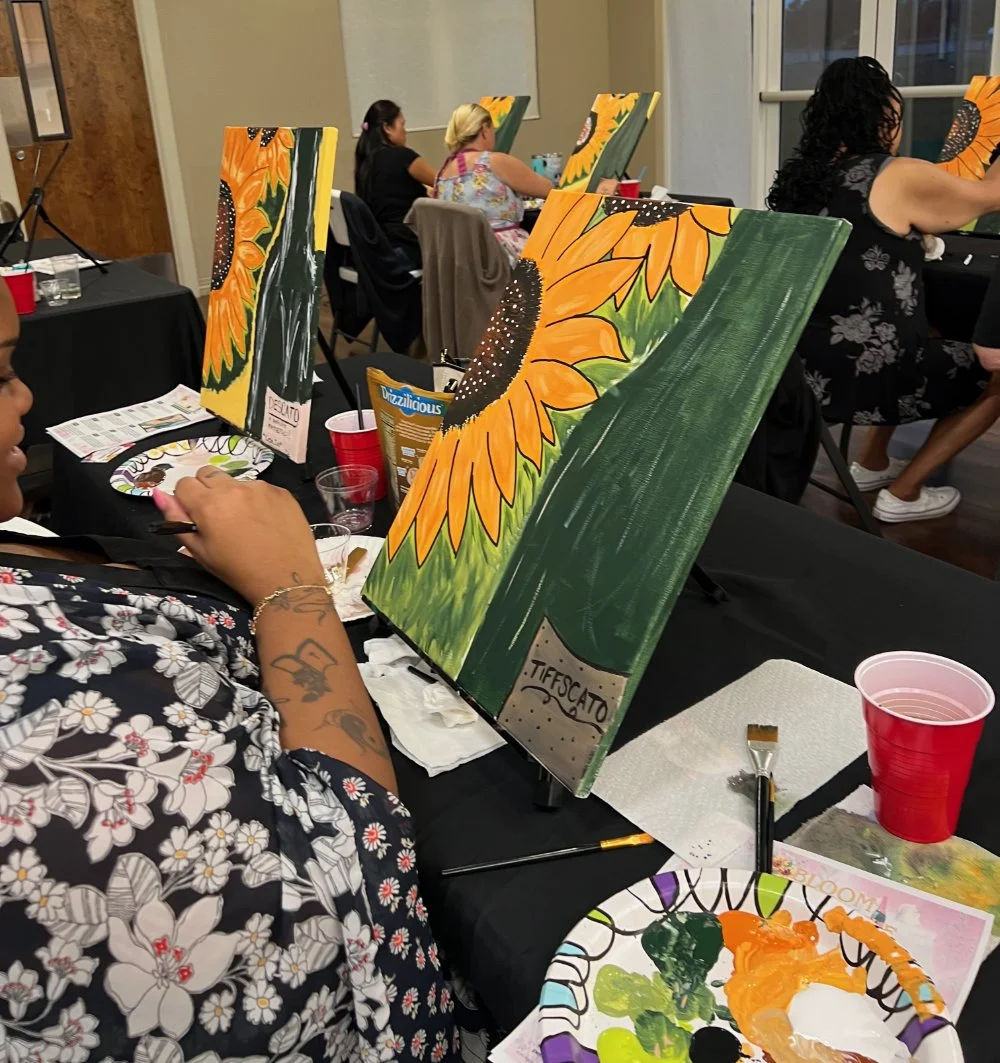 People participating in a painting class, painting sunflower-themed canvases. Art supplies and drinks are on the table.