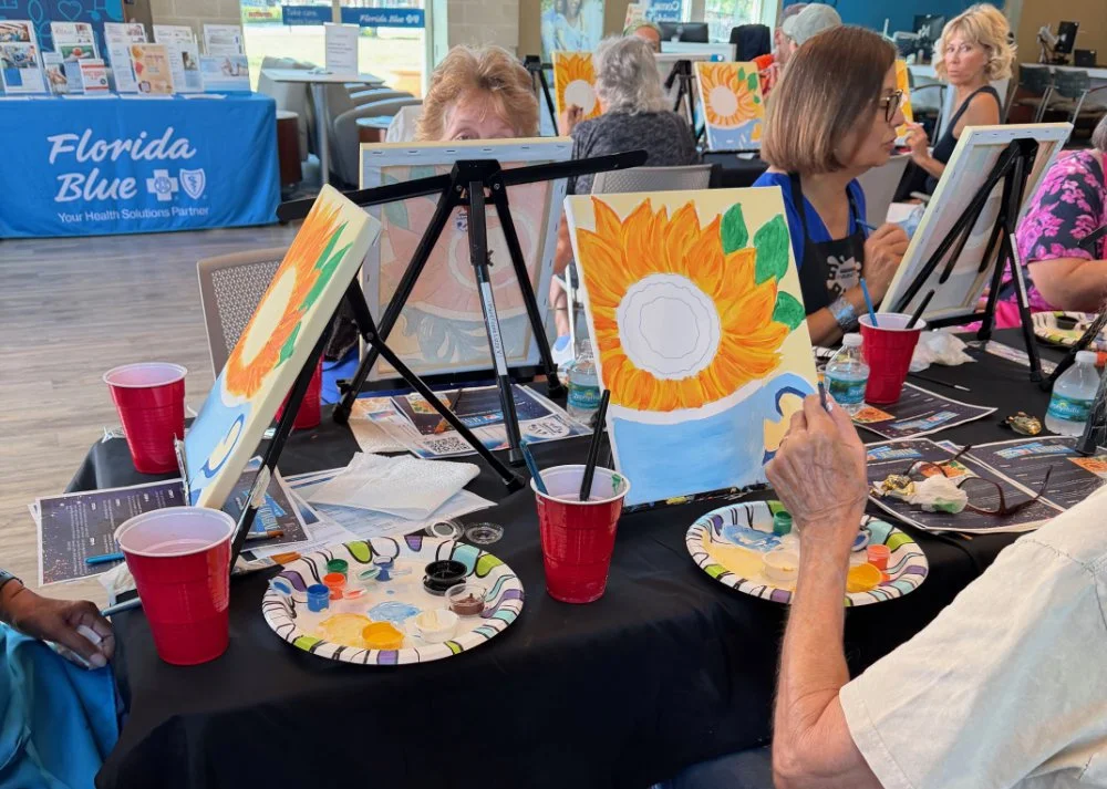 People participating in a paint and sip class, painting sunflowers on canvases. There are red cups, water bottles, and painting supplies on the table.
