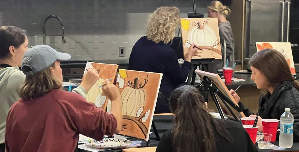 Group of people participating in a pumpkin painting class in a kitchen setting, painting pumpkins on canvases.