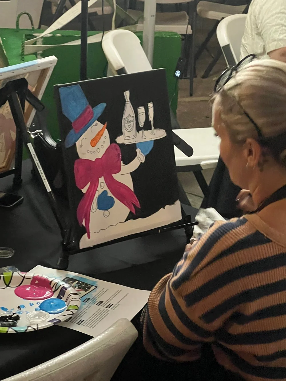 A person painting a snowman wearing a blue hat with a red band and blue mittens, holding a tray with a bottle and two glasses.