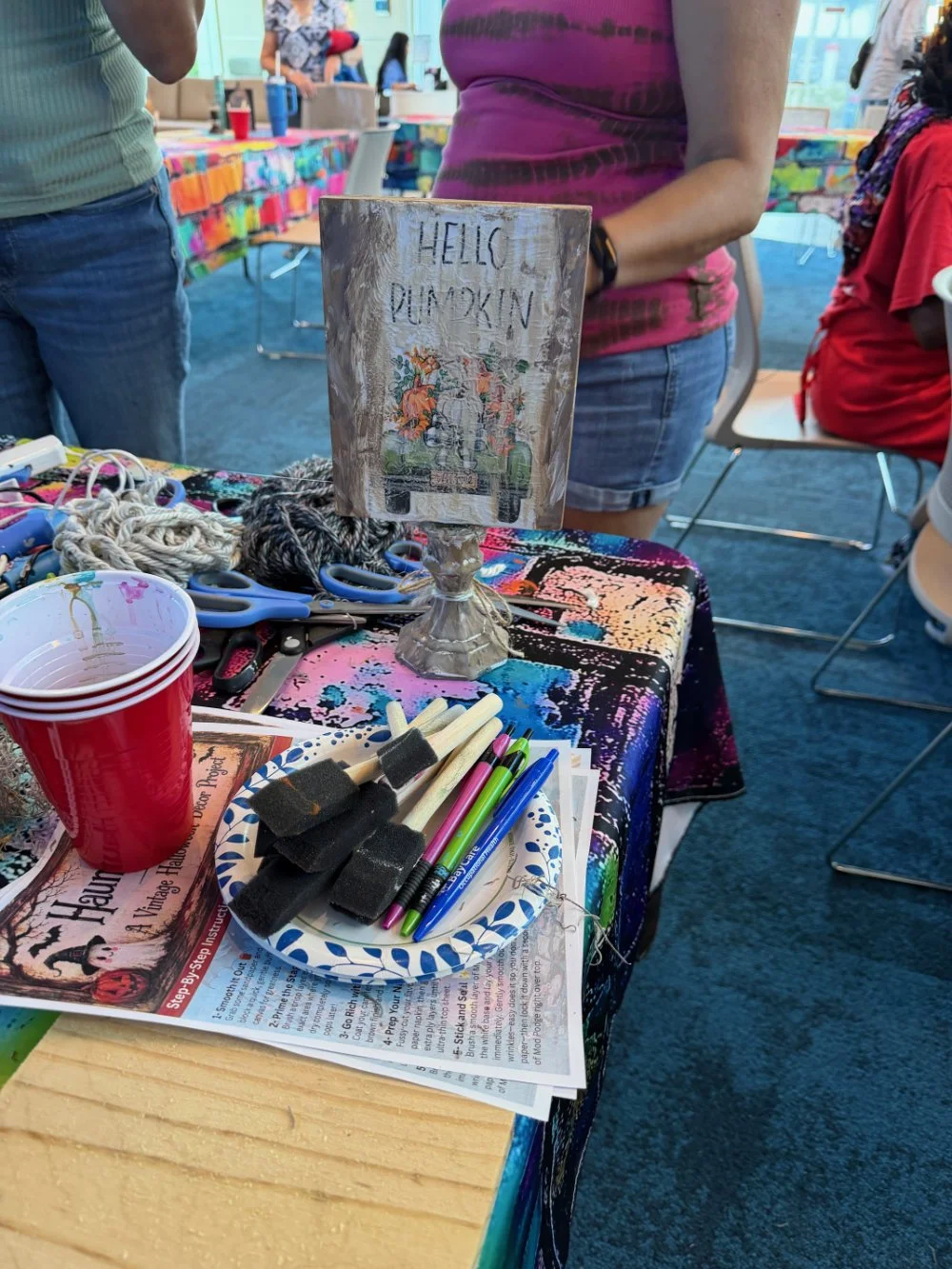 A table at a craft event with scissors, markers, foam brushes, yarn, and a sign that says 'Hello Pumpkin' with a fall-themed illustration.