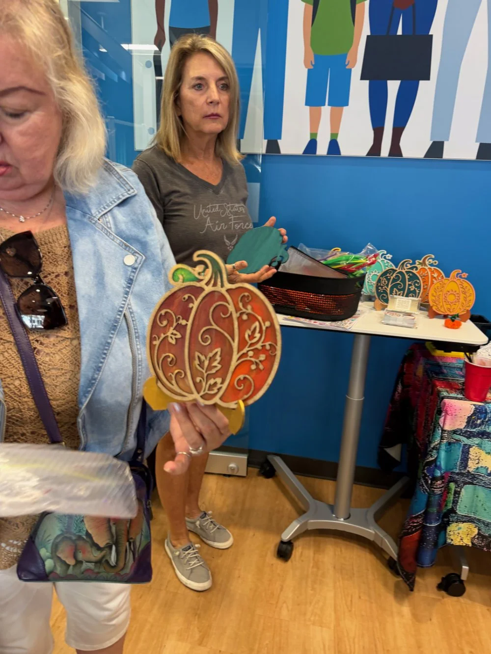 A woman holding a decorative pumpkin-shaped wooden craft with swirling patterns, standing near a table with other craft items, in an indoor setting.