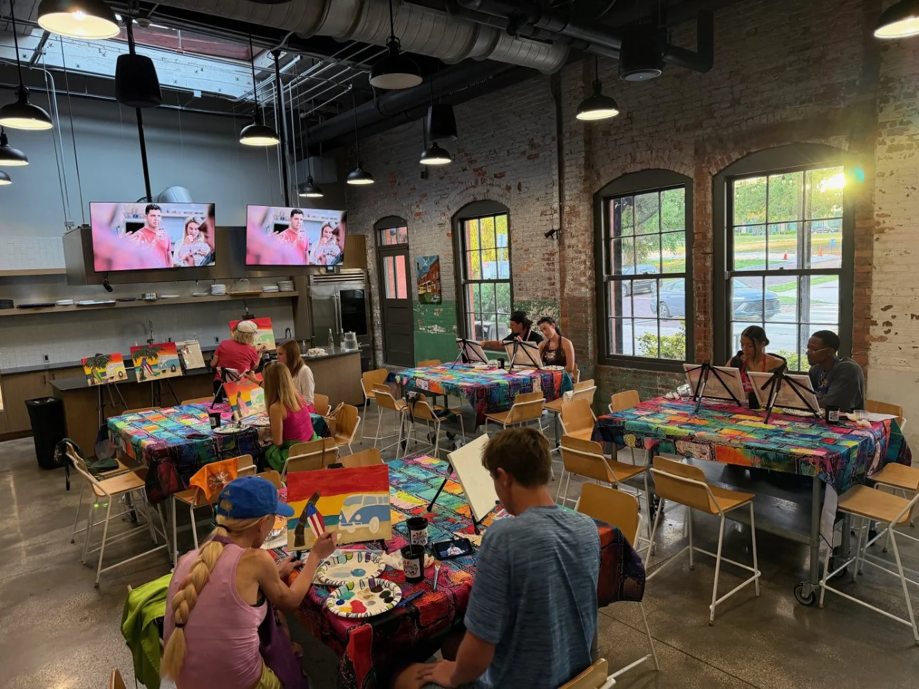 People participating in a painting class inside a spacious industrial-style room with large windows, colorful tablecloths, and paintings on display.