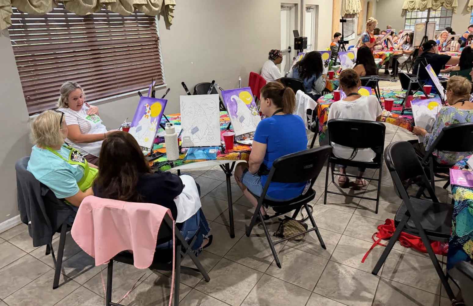 A group of people participating in a painting class at a restaurant. The tables are covered with colorful tablecloths, and the participants are working on canvas paintings featuring a moon and other designs. Some paintings are still in progress, and 