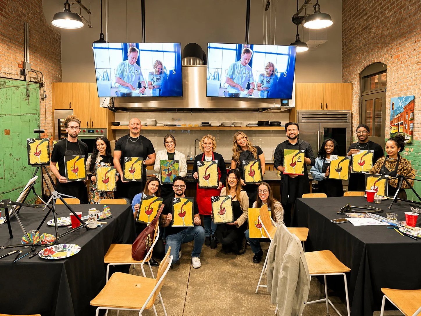 A group of people at a painting class in a studio, holding up their finished paintings of a wine glass with a cherry, while smiling at the camera. There are tables with art supplies and empty chairs, and two large screens displaying a video tutorial in the background.