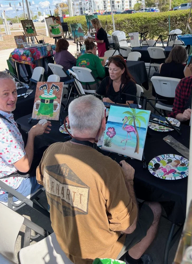A group of people participating in a painting event outdoors under a tent. Some are painting pictures of tropical scenes, including a flamingo and a palm tree, while others are observing or waiting for their turn. The setting appears festive and casu