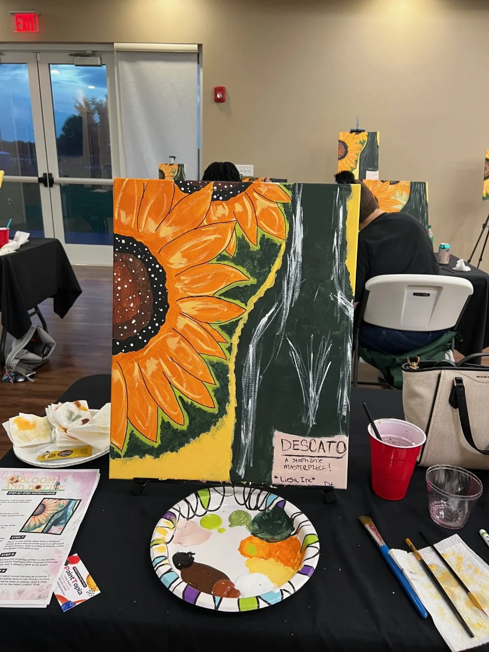 Painting of a sunflower and a vase on an easel at a painting class, with painting supplies on the table.