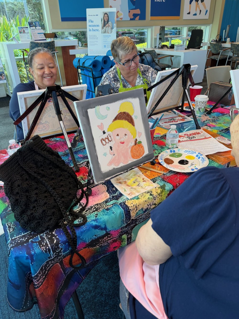 Two women sitting at a decorated table, painting pictures on small canvases set on easels. One woman is smiling, and the other is focused on her painting. The table has art supplies, water bottles, cups, and a colorful tablecloth. There's a small fra