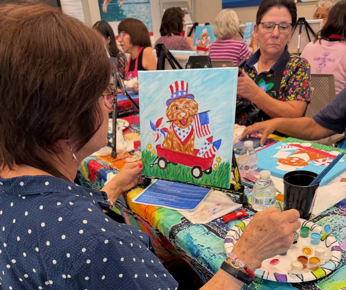 A group of women participating in a painting event, with a painting of a dog dressed as Uncle Sam in a red wagon as the focal point.