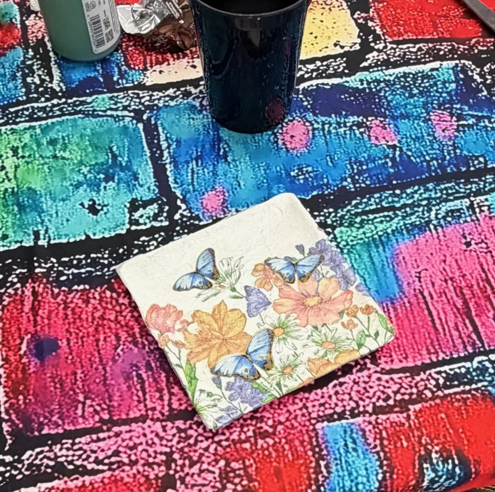 Colorful tablecloth with a floral napkin featuring butterflies, a black cup, and some cans in the background.