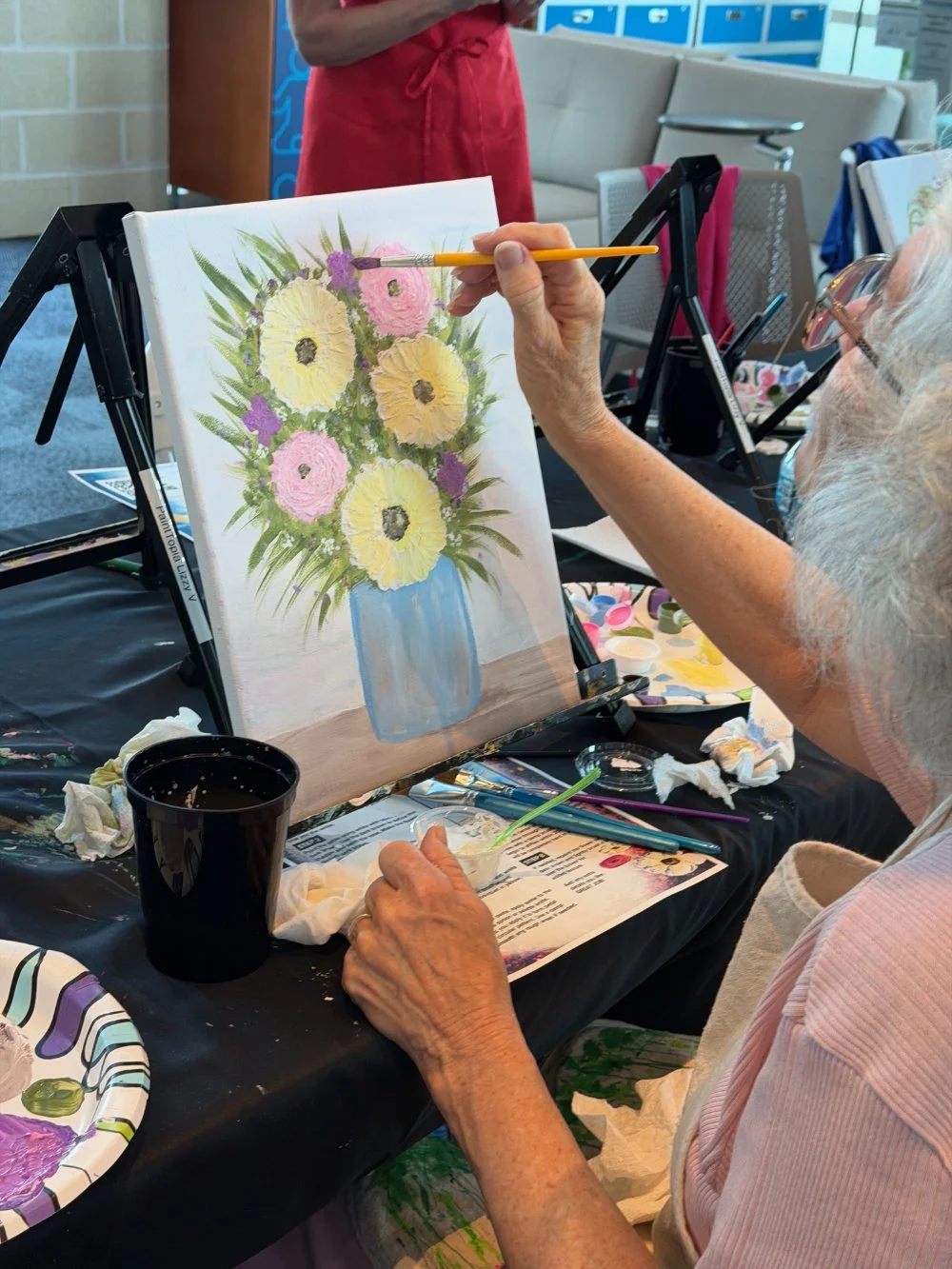 An elderly woman painting a floral arrangement in a vase on an easel at an art class.