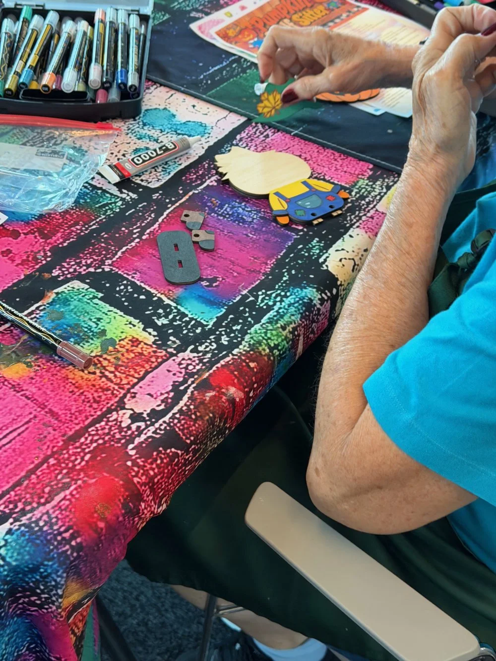 A person with gray hair working on a craft project at a colorful table, surrounded by supplies such as markers, small wooden pieces, and a piece with a cartoon design.