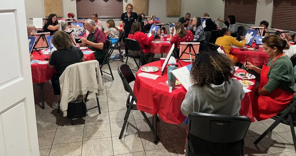 People sitting at tables with easels, painting during a group art class or workshop, in a room with wooden blinds and decorated with red-themed table settings.