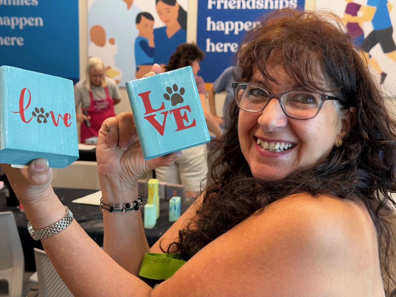 A woman with glasses and curly hair smiling and holding three wooden blocks that spell out love in a heart-shaped design, with a background of blue wall and other people at a table.