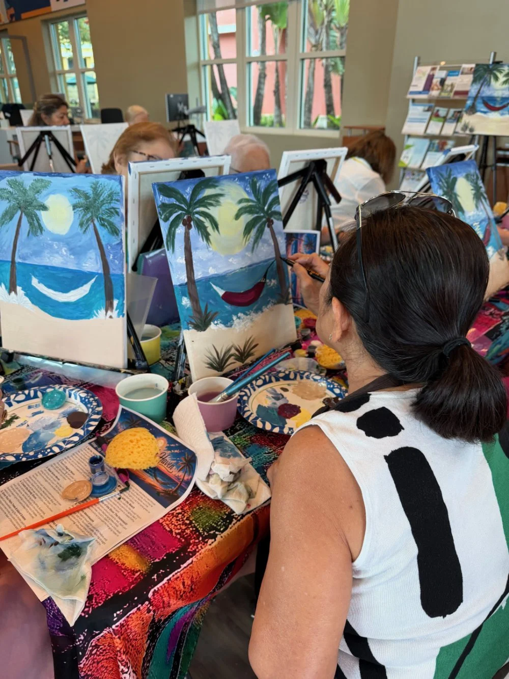 People participating in a painting class, creating tropical beach scenes with palm trees and a sun, at a table covered with art supplies and painted canvases.