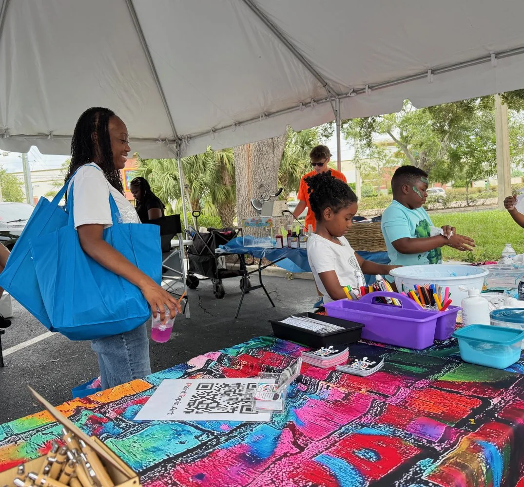 People at a booth with arts and crafts supplies under a white canopy tent in a park-like setting.