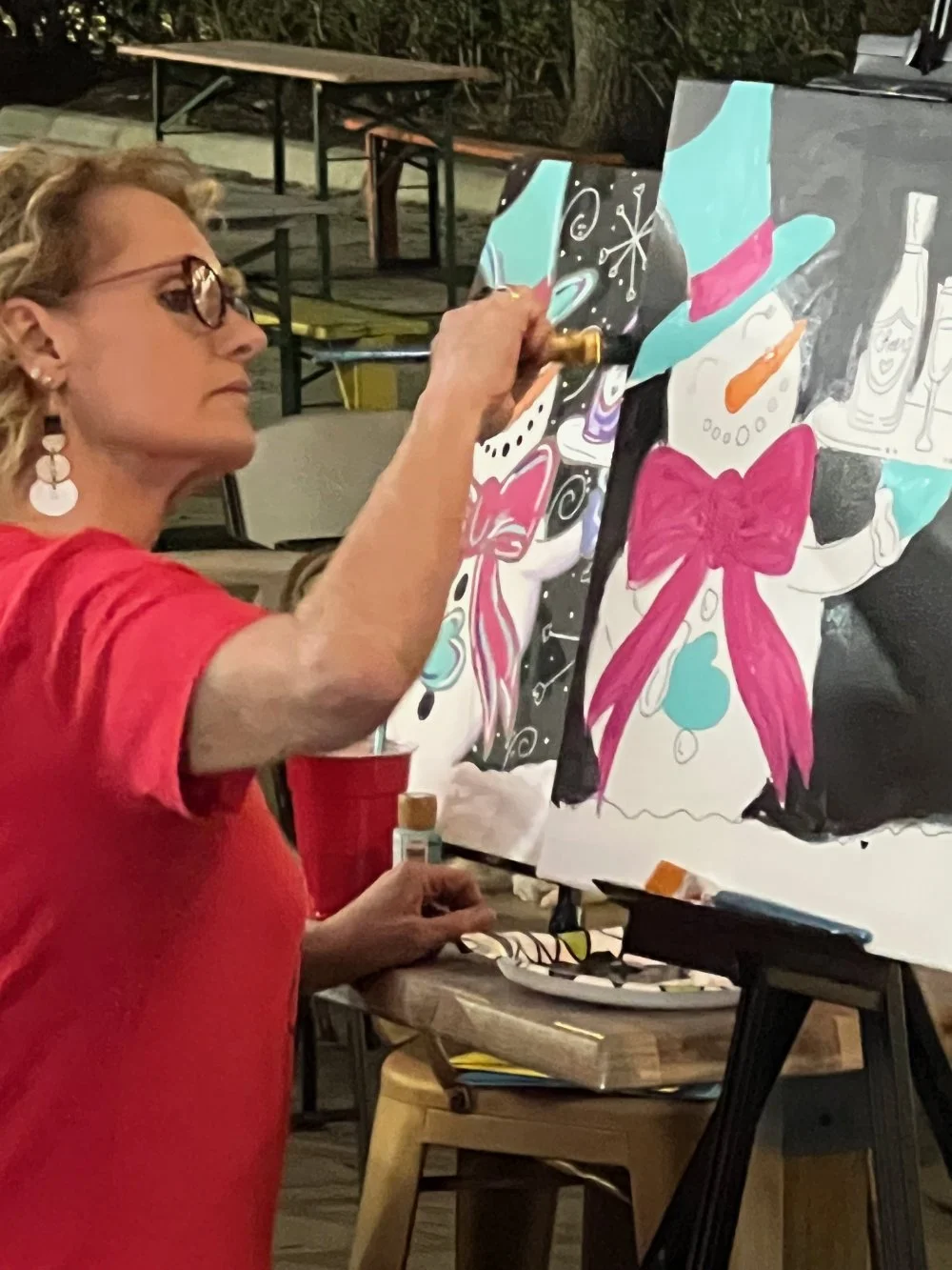 A woman wearing a red shirt and glasses is painting a holiday-themed design on a canvas, featuring a large pink bow, a snowman, and festive decorations.
