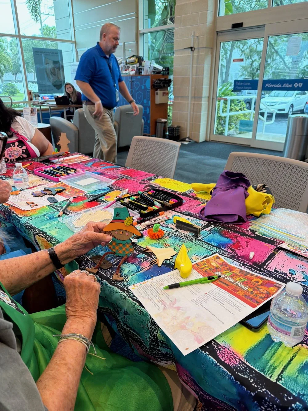 Elderly woman holding a colorful paper craft while sitting at a table with arts and crafts supplies, including markers, paper, and a water bottle, at a community center with large windows and colorful tablecloth.