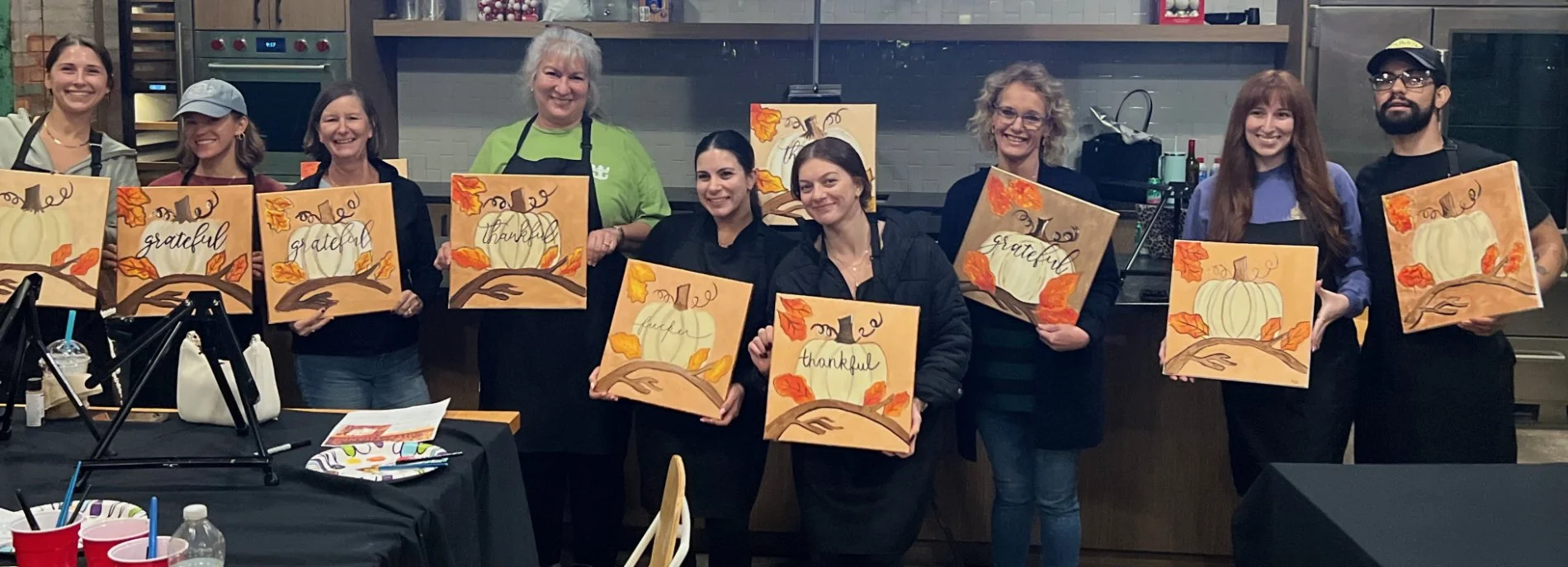 A group of nine people standing indoors, each holding a fall-themed canvas painting with a pumpkin, leaves, and words like 'thankful,' 'grateful,' and 'blessed.' They are smiling and appear to have participated in a painting class.