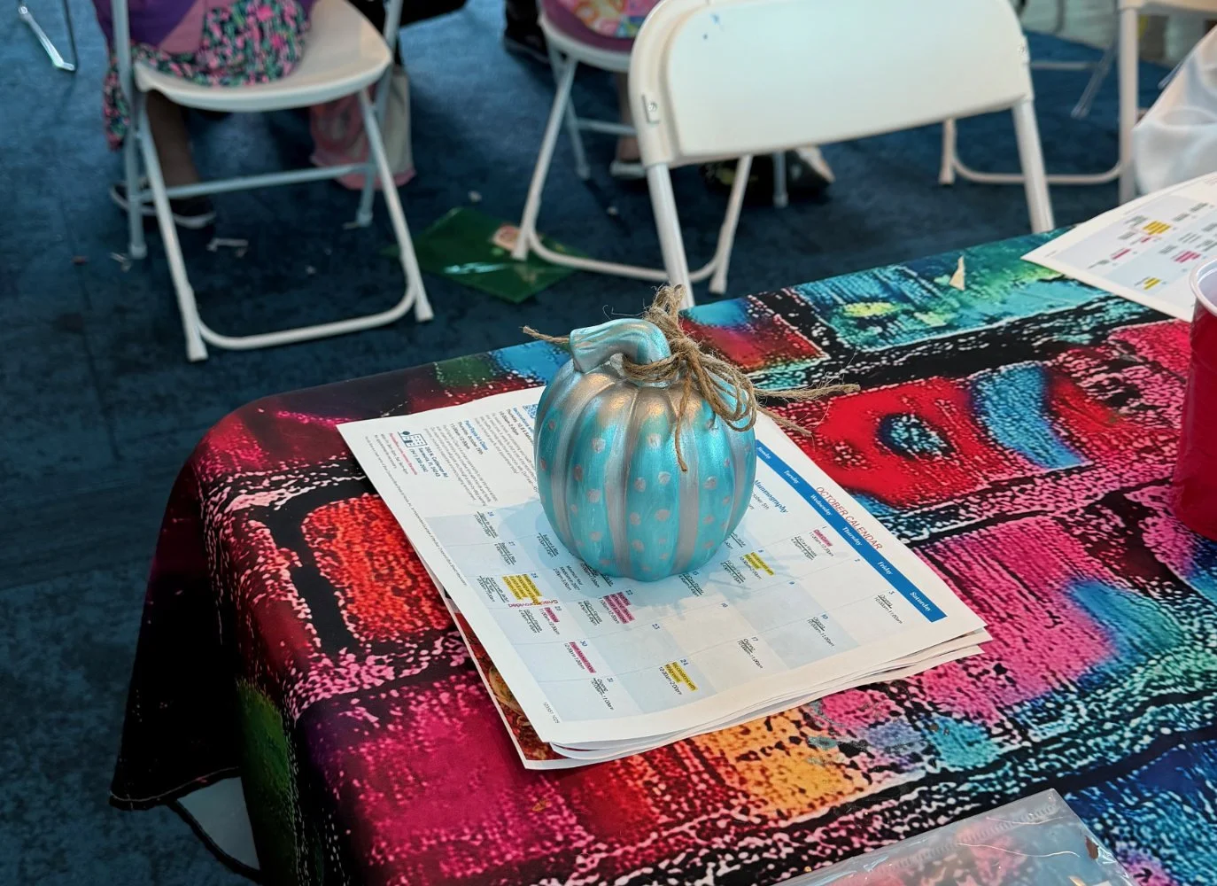 Table with a colorful tablecloth, a ceramic pumpkin with a twine bow on top, and papers underneath, in a setting with white chairs around.