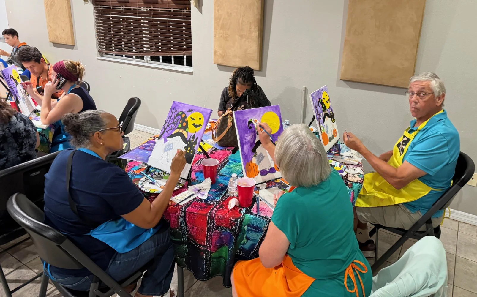 People painting Halloween-themed pictures at a craft party, with decorations including pumpkins and a full moon.
