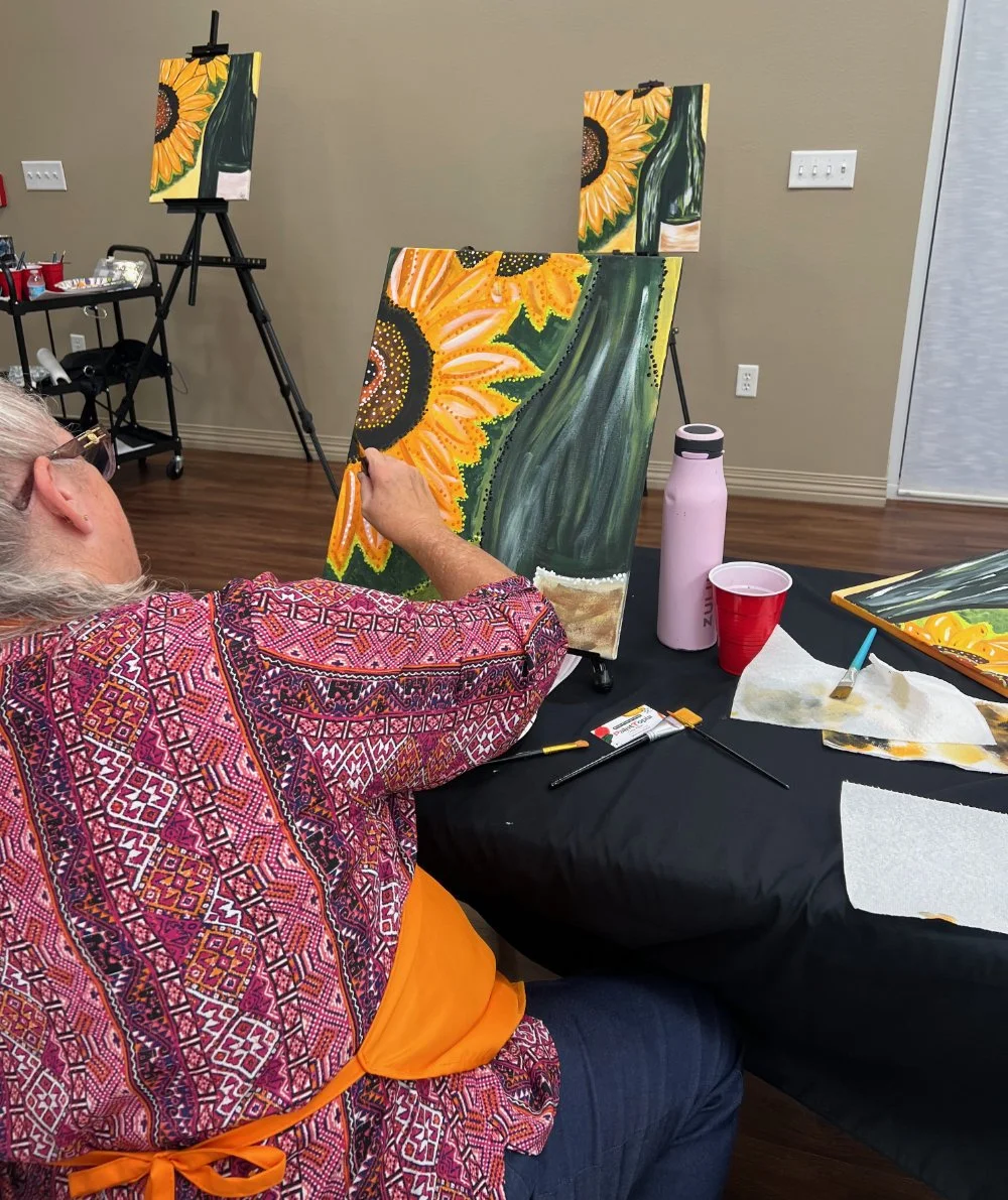 A woman painting a sunflower on a canvas at an art class. The painting depicts a large sunflower with bright yellow petals and a dark center. The woman is seated at a table with painting supplies, wearing a colorful patterned shirt and an orange apro