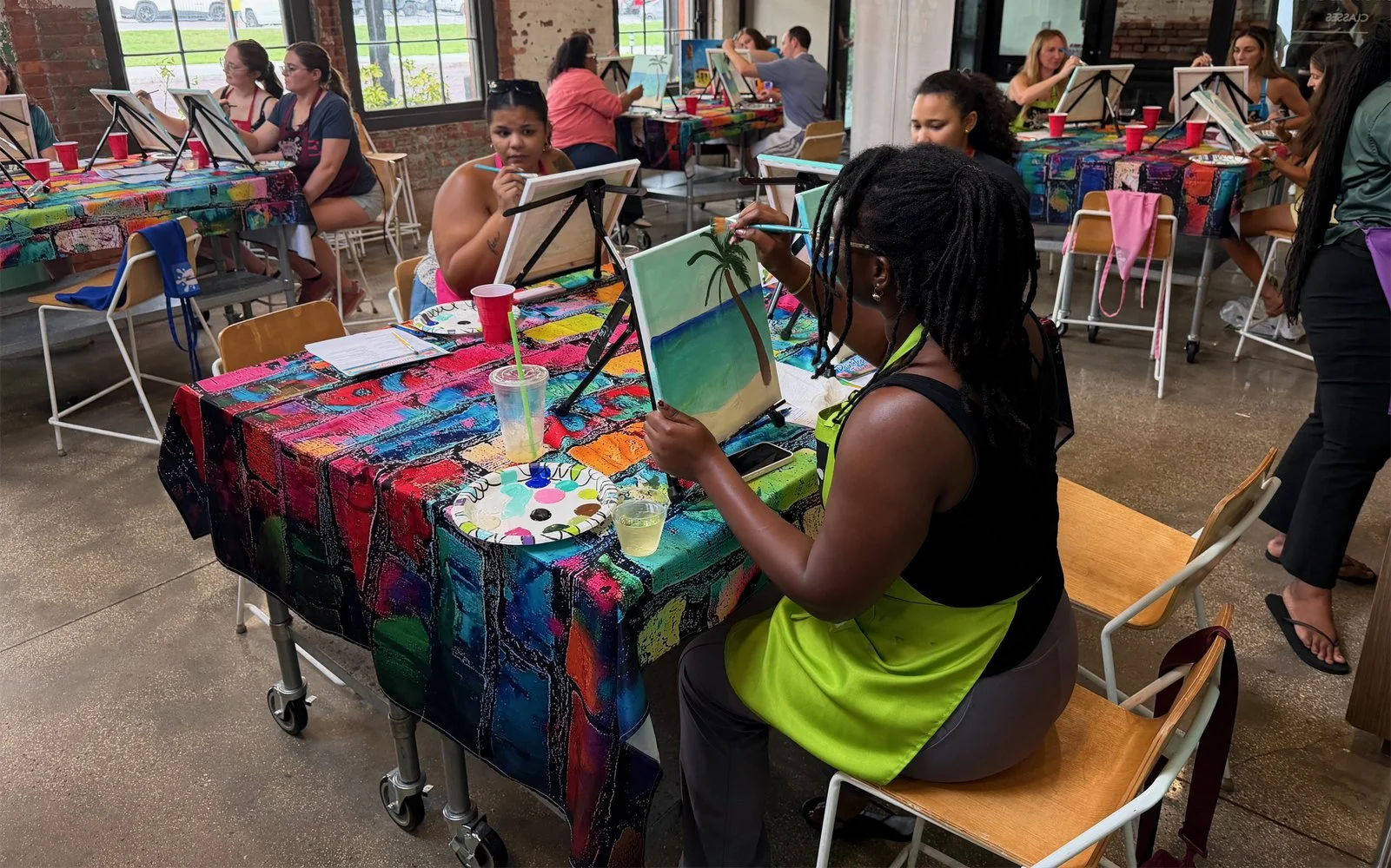 A group of people painting at tables covered with colorful, abstract tablecloths in an indoor space with large windows and exposed brick walls.