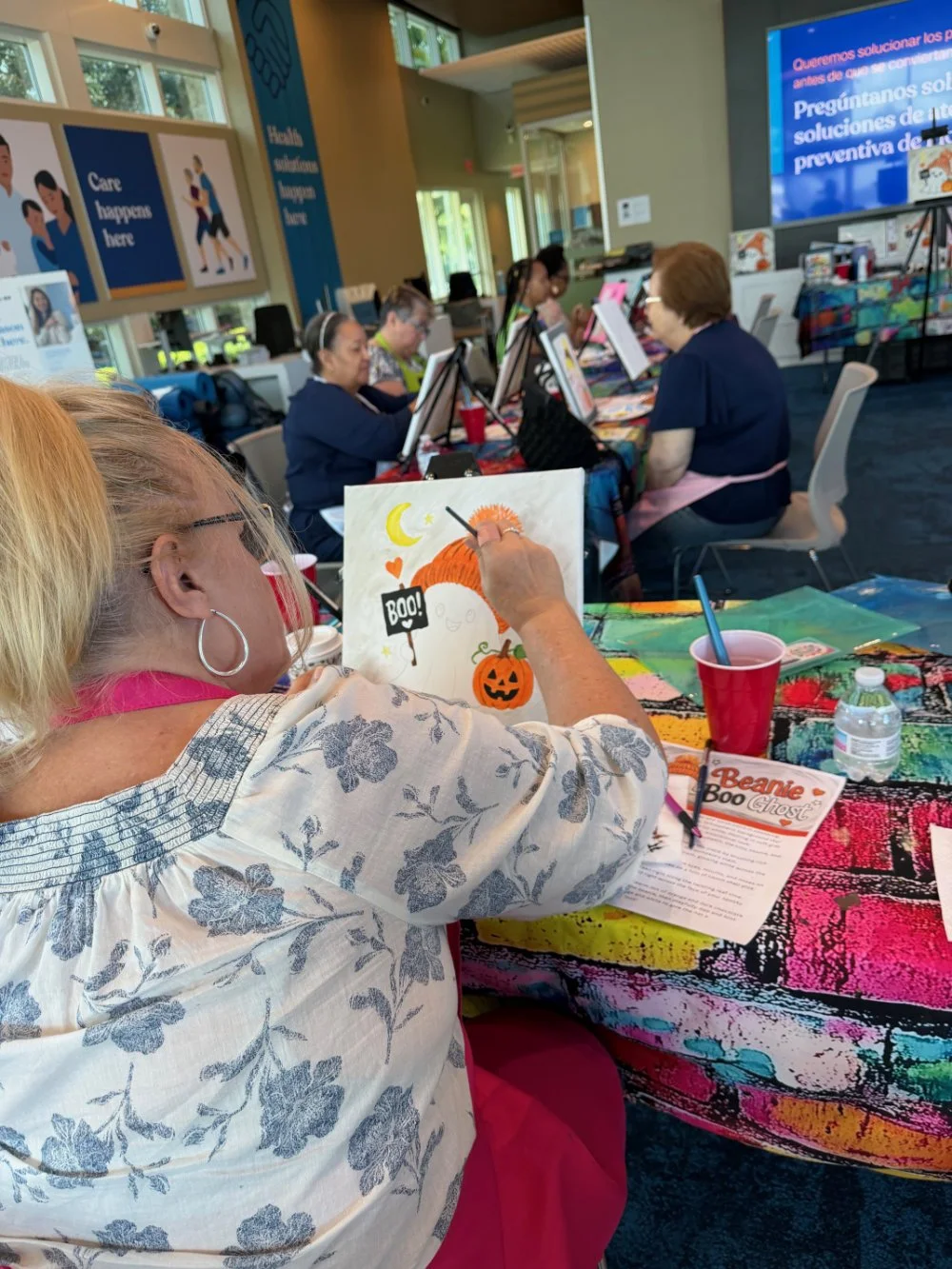 A group of women seated at a colorful table participating in a painting activity. The woman in the foreground is painting a Halloween-themed picture featuring a pumpkin, moon, and stars. In the background, a large screen and posters with motivational