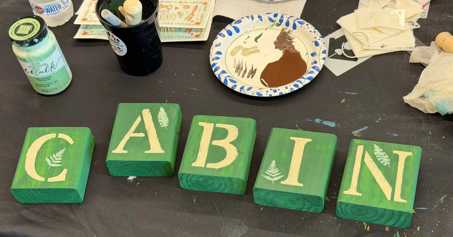 Colorful painted blocks spelling out C A N, with leaf designs on a black table alongside craft supplies including paint, paper plates, and a mug with paintbrushes.